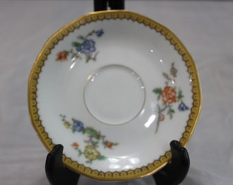 May include: A white porcelain saucer with a gold rim and floral pattern. The saucer has a small, round depression in the center.