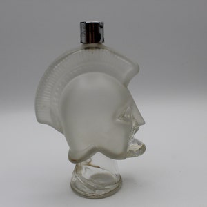 May include: A frosted glass bottle shaped like a Roman warrior's helmet with a silver cap. The bottle is clear glass with a textured surface.