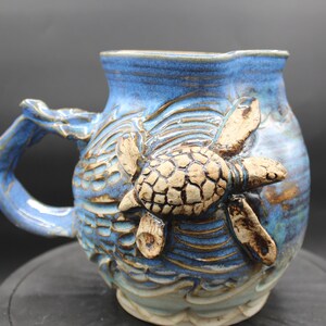 May include: Blue ceramic mug featuring a 3D sea turtle design. The turtle is brown and white and is swimming in waves. The mug has a blue glaze with brown accents.