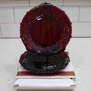 May include: Two deep red glass plates featuring a textured, embossed pattern along the scalloped edges and a central sunburst design. One plate is displayed upright on a black metal stand, while the second plate rests flat on its original box.