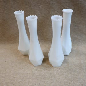 May include: Four white glass vases with a unique geometric base and ruffled top edge. The vases have a slender, elongated shape, and are arranged on a neutral-toned surface. These decorative vases are suitable for floral arrangements.