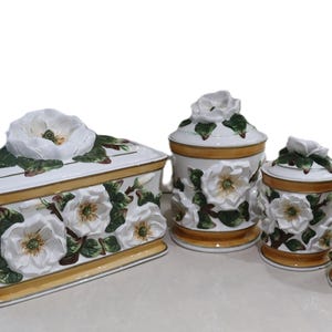 May include: A set of four ceramic kitchen canisters and a rectangular box. The white pieces are adorned with 3D white flower and green leaf designs. The set has gold trim and lids topped with flower accents.