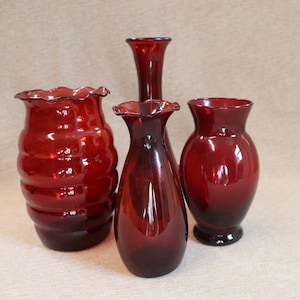 Op de afbeelding: Vier robijnrode glazen vazen in verschillende vormen en maten. Eén vaas heeft een gerimpelde rand, een andere heeft een bolvormige vorm en de andere zijn meer gestroomlijnd. De vazen zijn geplaatst op een neutrale achtergrond.