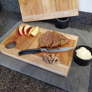 May include: A wooden cutting board with a handle, divided into two sections. One section has slices of apple, the other has bread, pistachios, and a small bowl of butter.