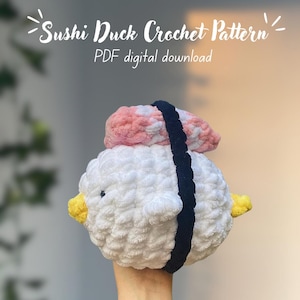 Puede incluir: Un juguete amigurumi de pato sushi tejido a crochet. El pato es blanco con una cobertura de sushi rosa y blanca y una envoltura de alga negra. El juguete está siendo sostenido por una mano.