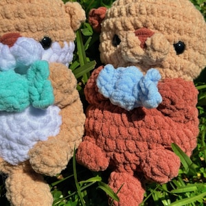 May include: Two crocheted stuffed sea otters holding blue and green fish. The otters are brown and white with black eyes. The otters are sitting on a bed of green grass.