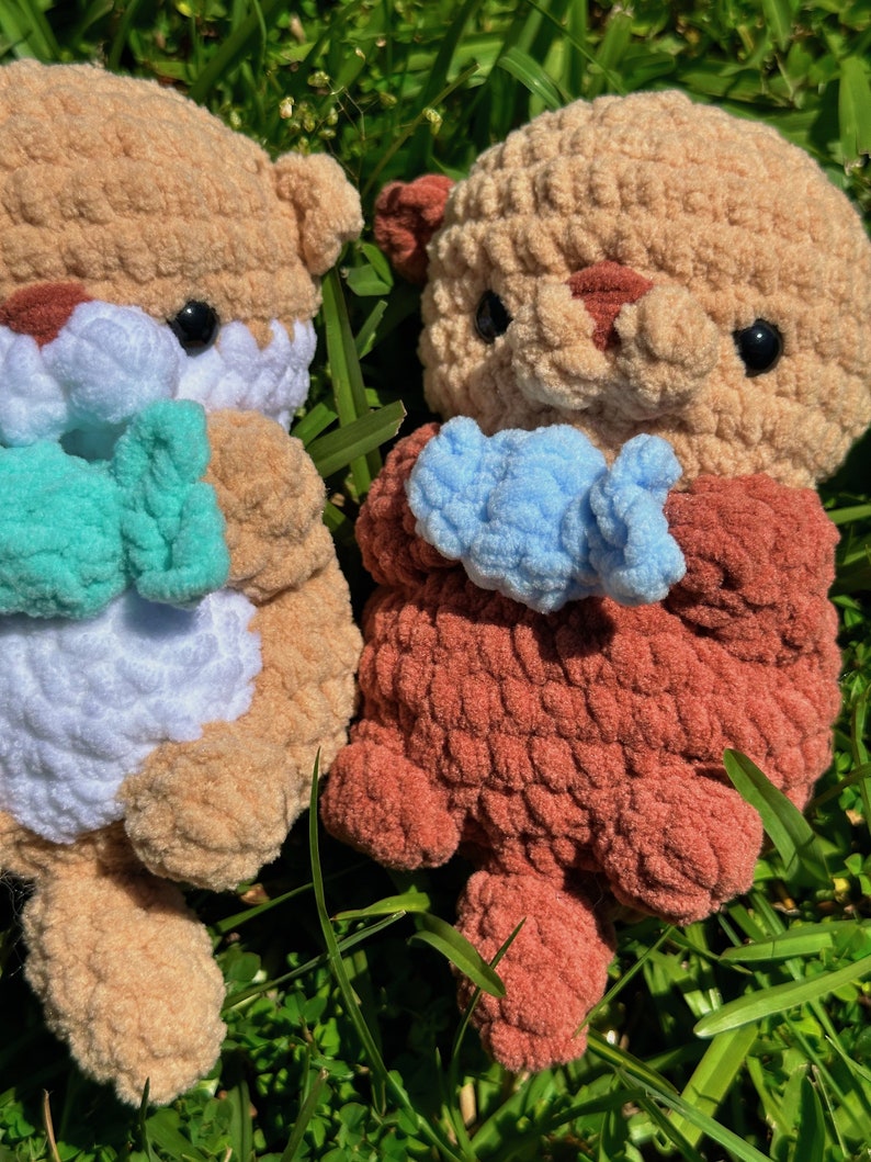 May include: Two crocheted stuffed sea otters holding blue and green fish. The otters are brown and white with black eyes. The otters are sitting on a bed of green grass.