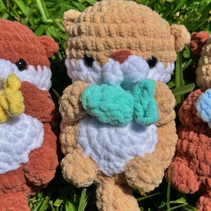 May include: Three crocheted stuffed animals in the shape of otters. The otter in the centre is brown and white and is holding a teal blue crocheted fish. The otters on either side are brown and white and are holding yellow and blue crocheted fish.