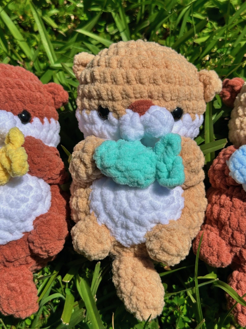 May include: Three crocheted stuffed animals in the shape of otters. The otter in the centre is brown and white and is holding a teal blue crocheted fish. The otters on either side are brown and white and are holding yellow and blue crocheted fish.