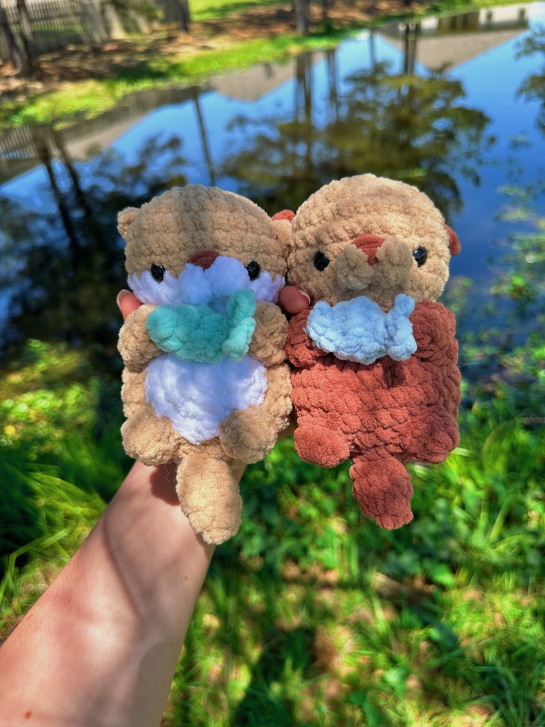 May include: Two crocheted stuffed sea otter plushies holding blue and green fish. The otters are brown and white and are being held in a hand.