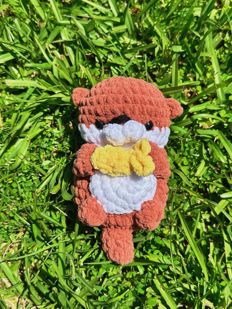 May include: A crocheted plush toy otter in shades of brown and white, holding a yellow crocheted fish in its paws. The otter is sitting on a bed of green grass.