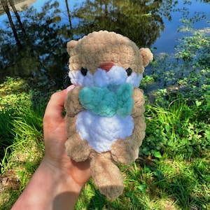 May include: A crocheted plush otter toy holding a green crocheted fish. The otter is brown and white with black eyes and a brown nose. The toy is being held by a hand in front of a green grassy background with a pond in the distance.