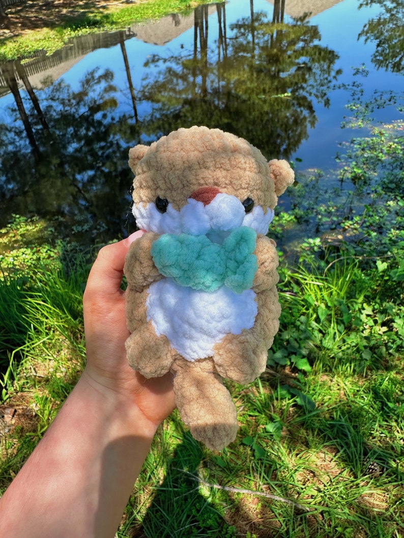 May include: A crocheted plush otter toy holding a green crocheted fish. The otter is brown and white with black eyes and a brown nose. The toy is being held by a hand in front of a green grassy background with a pond in the distance.
