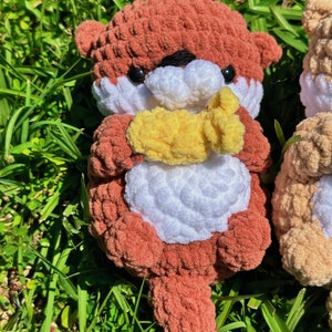 May include: A crocheted plush toy otter in shades of brown and white, holding a yellow crocheted fish in its paws. The otter is sitting on a bed of green grass.