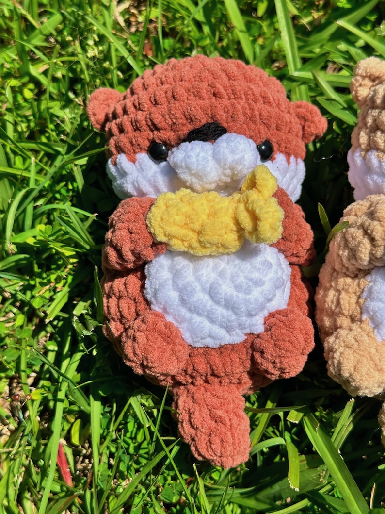 May include: A crocheted plush toy otter in shades of brown and white, holding a yellow crocheted fish in its paws. The otter is sitting on a bed of green grass.