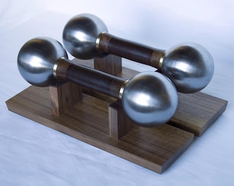 May include: Two dumbbells with polished metal spheres and dark wood handles, accented with brass rings. The dumbbells rest on a wooden stand with a natural finish, showcasing a minimalist design. Ideal for home fitness and weight training.