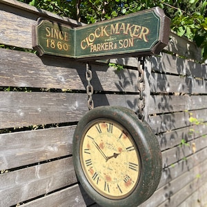 May include: A double-sided wall-mounted clock with a dark green frame and a distressed cream face featuring Roman numerals. The sign above reads "SINCE 1860" and "CLOCK MAKER PARKER & SON" in gold lettering, suspended by two metal chains.