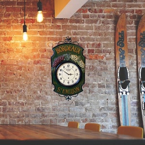 May include: A rustic wooden dining table with six yellow chairs. A vintage clock with a green face and gold accents hangs on a brick wall. The clock features the text "Bordeaux St. Emilion". Two wooden skis are leaning against the wall.