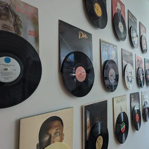 Puede incluir: Una colección de discos de vinilo exhibidos en una pared blanca. Los discos están en sus fundas, mostrando portadas de álbumes. Los discos negros tienen etiquetas coloridas, creando una estética vintage.