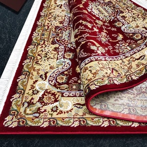 Puede incluir: Una alfombra roja con un patr&oacute;n floral crema y dorado. La alfombra tiene un borde de flecos y est&aacute; enrollada.
