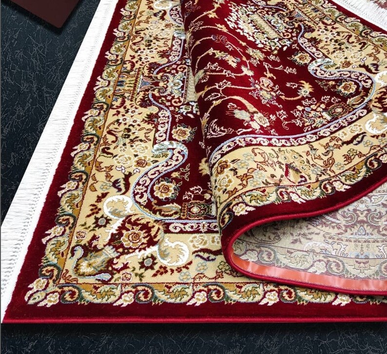 Puede incluir: Una alfombra roja con un patr&oacute;n floral crema y dorado. La alfombra tiene un borde de flecos y est&aacute; enrollada.