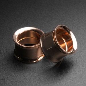 Könnte beinhalten: Zwei Ohrdehner aus Roségold, einer größer als der andere, werden auf einer schwarzen Oberfläche gezeigt. Die Dehnungen sind glatt und haben eine glänzende Oberfläche.