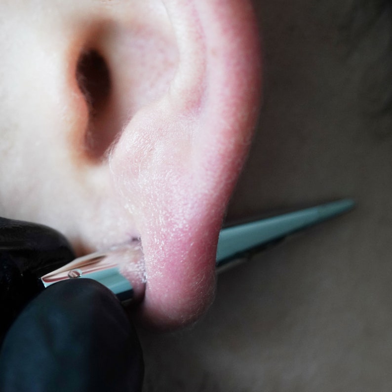 May include: Close-up of a person's ear with a piercing being inserted using a tool.