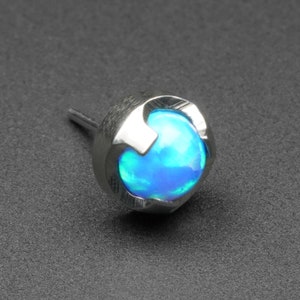 May include: A single, silver-colored stud earring with a bright blue opal-like stone. The stone is round and set within a decorative, open-work bezel. The earring post is visible.