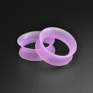 May include: Two purple silicone ear gauges, one slightly larger than the other, are shown on a black background.