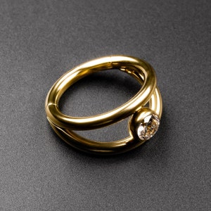 May include: A gold ring with a single, round, clear diamond set in the center. The ring is made of two bands that are intertwined.