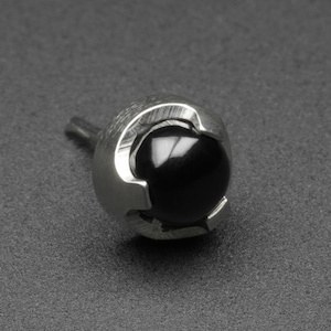 May include: A silver-colored, round stud earring with a black, spherical center. The earring has a textured, metallic design around the black sphere and a small post for insertion. The background is a dark gray.