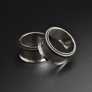 May include: Two silver metal ear gauges with black rubber rings. The gauges are stacked on top of each other.