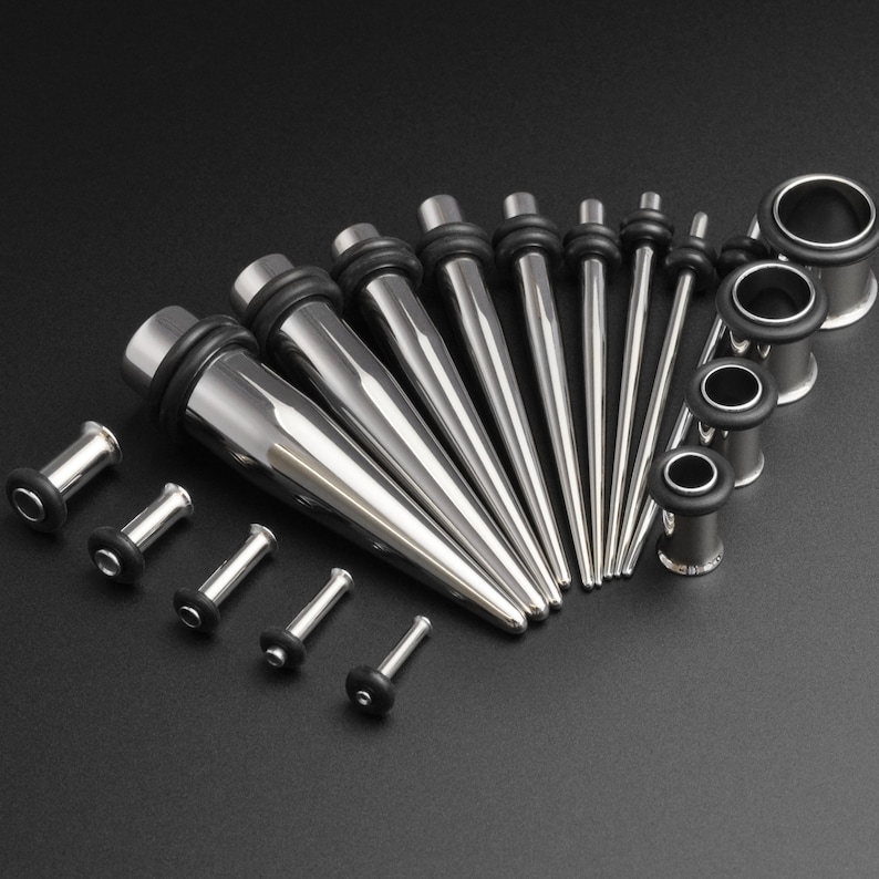 May include: A set of 14 stainless steel ear gauges, including tapers and tunnels, in various sizes. The gauges are arranged in a fan shape on a black background.