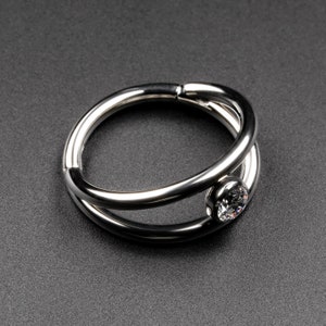 May include: A silver ring featuring a clear, round gemstone. The ring's design incorporates two intertwined bands that intersect. The gemstone is set within a bezel. The ring is displayed against a dark background.