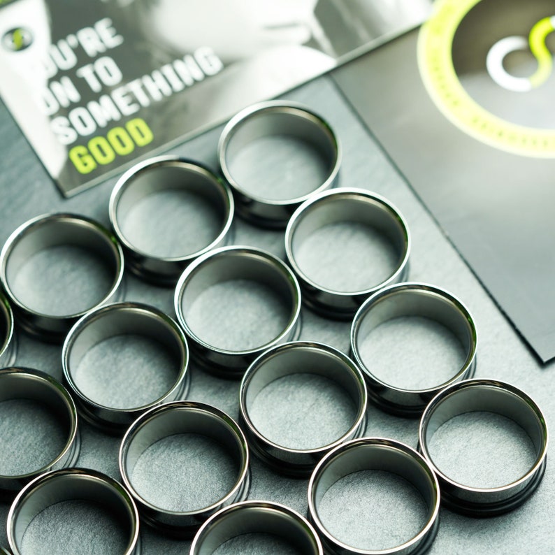 May include: A set of 12 silver-colored metal ear gauges, arranged in a grid pattern on a black surface. The gauges are of varying sizes, with the largest gauge in the center of the grid.