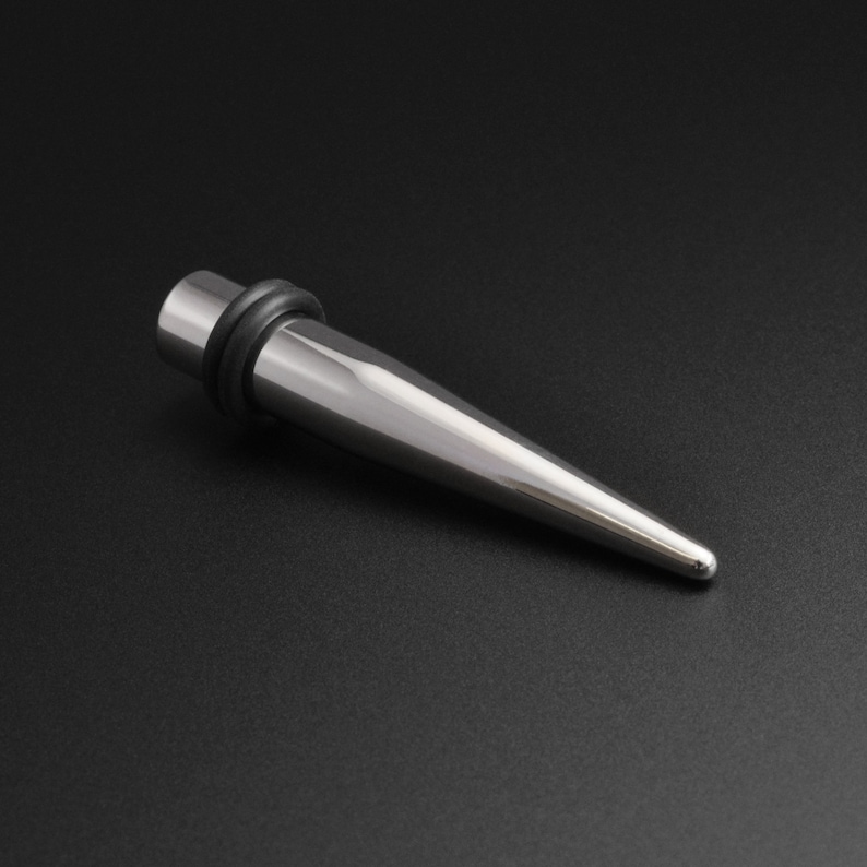 May include: A silver metal taper with a black rubber o-ring. The taper is pointed at one end and has a flat surface at the other end.