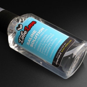 Puede incluir: Una botella transparente de solución salina "Lobe Love" para piercings. La etiqueta es azul claro con texto blanco y rojo, con instrucciones de uso. La botella tiene una tapa negra y está sobre una superficie negra.