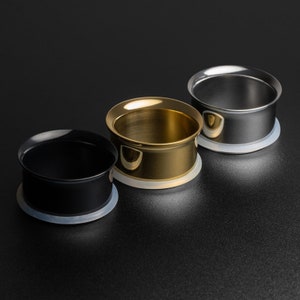 Puede incluir: Tres expansores de oreja, uno negro, uno dorado y uno plateado, con un respaldo de silicona blanco. Los expansores están dispuestos en una fila de izquierda a derecha.