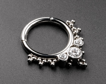 Könnte beinhalten: Ein silberner, klappbarer Septumring mit drei klaren, runden Edelsteinen und kleinen Perlenverzierungen. Der kreisförmige Körperschmuck wird vor einem dunkelgrauen Hintergrund gezeigt.