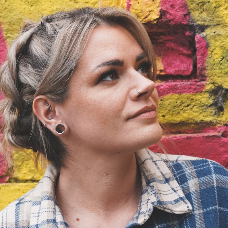 May include: A woman with blonde hair and a plaid shirt looks up towards the right. She has multiple piercings in her left ear, including a large hoop.
