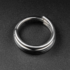 May include: A shiny, silver-colored metal ring with a triple-band design. The ring is circular and appears to be made of a smooth, polished material. The background is a dark, textured surface.