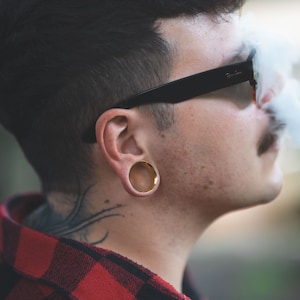 Peut inclure: Un homme aux cheveux noirs courts portant des lunettes de soleil &agrave; monture noire et une chemise &agrave; carreaux rouge et noir. Il porte une boucle d'oreille dor&eacute;e &agrave; l'oreille droite.