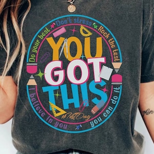 May include: Dark gray t-shirt with a colorful graphic that says "YOU GOT THIS" in large letters. The design includes school supplies and the phrases "Do your best," "Rock the test," and "I believe in you."