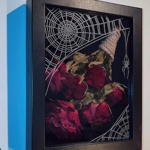May include: A framed artwork with dried red roses, a spider web design, and a small spider. The roses are tied with twine. The artwork is in a black shadow box frame, creating a gothic or Halloween theme.