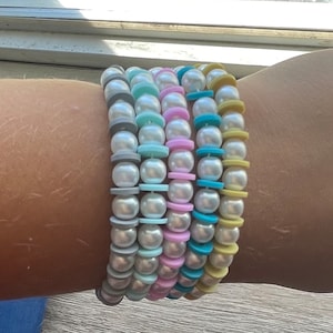 May include: A collection of six beaded bracelets. Each bracelet features white faux pearls and colorful spacers in shades of gray, blue, pink, and yellow. The bracelets are stacked on top of each other.