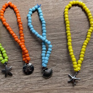 May include: Five beaded bracelets in shades of green, orange, light blue, yellow, and blue. Each bracelet features a charm with a sea-themed design, including a starfish, sand dollar, and seashell. The bracelets are made with small, round beads.