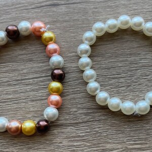 May include: Two beaded bracelets. One bracelet features a mix of white, brown, gold, and peach-coloured beads. The other bracelet is made of white beads. Both bracelets are on a wooden surface.