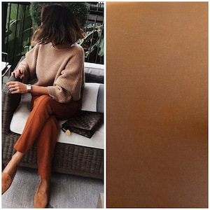 Puede incluir: Una mujer con un suéter marrón claro y pantalones naranja se sienta en una silla de mimbre. Lleva un embrague de cuero marrón. El fondo es una imagen borrosa de un patio.