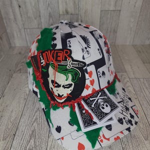 May include: White baseball cap with a playing card pattern and a Joker patch. The patch features the Joker's face with green hair and red accents. The word "JOKER" is displayed above the patch.