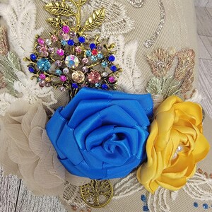 May include: A beige baseball cap adorned with a gold-toned tree branch and a jeweled tree brooch. Features include a blue satin rose, a yellow satin rose, and delicate lace and glitter accents. The cap is embellished with small blue and clear rhinestones.